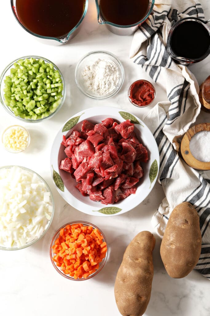 Ingredients to make beef stew in the slow Coker in bowls on a marble surface with a black and white striped linen