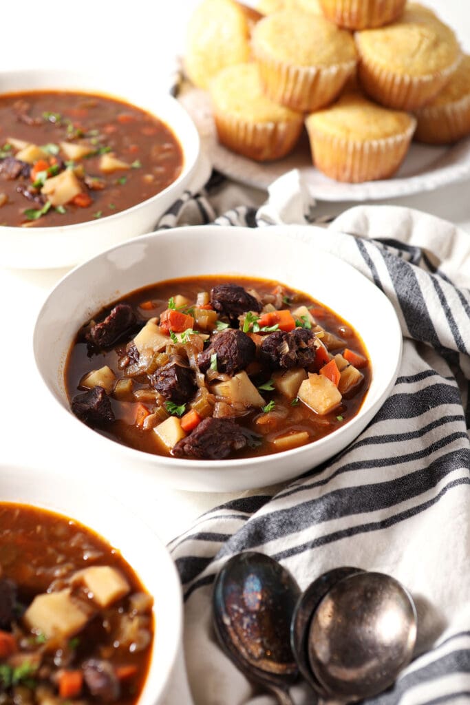 Three bowls of beef stew on marble with a striped linen and a plate of cornbread muffins