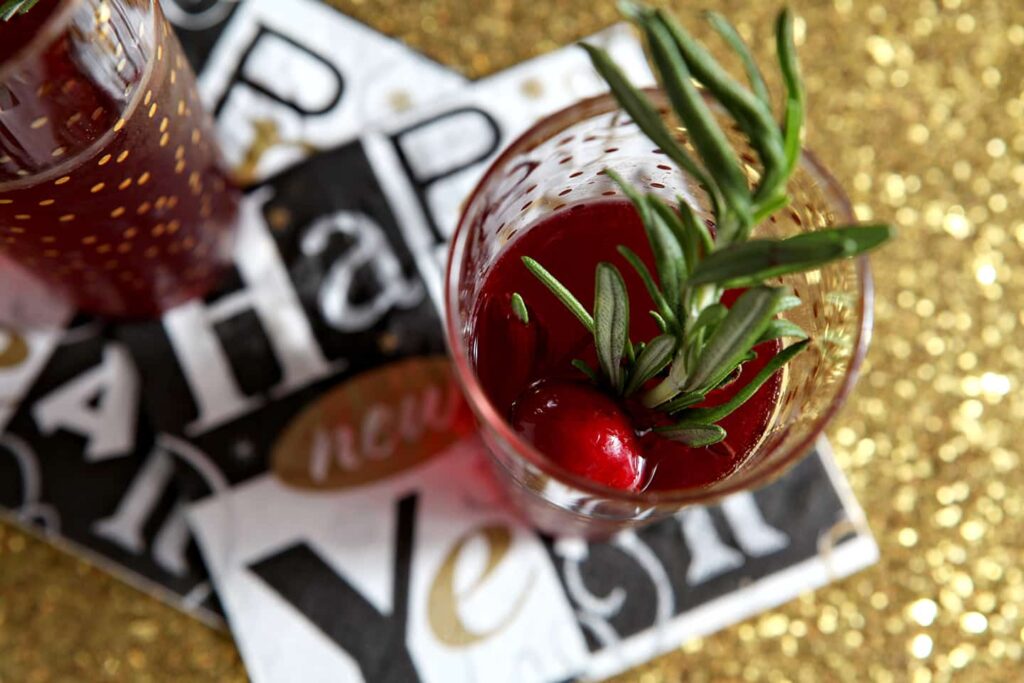 Two champagne flutes holding red liquid and fresh rosemary from above on a gold glitter platter