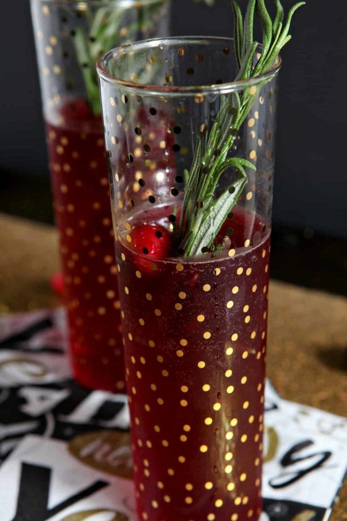 Close up of two champagne flutes holding red drinks with fresh cranberry and rosemary garnishes