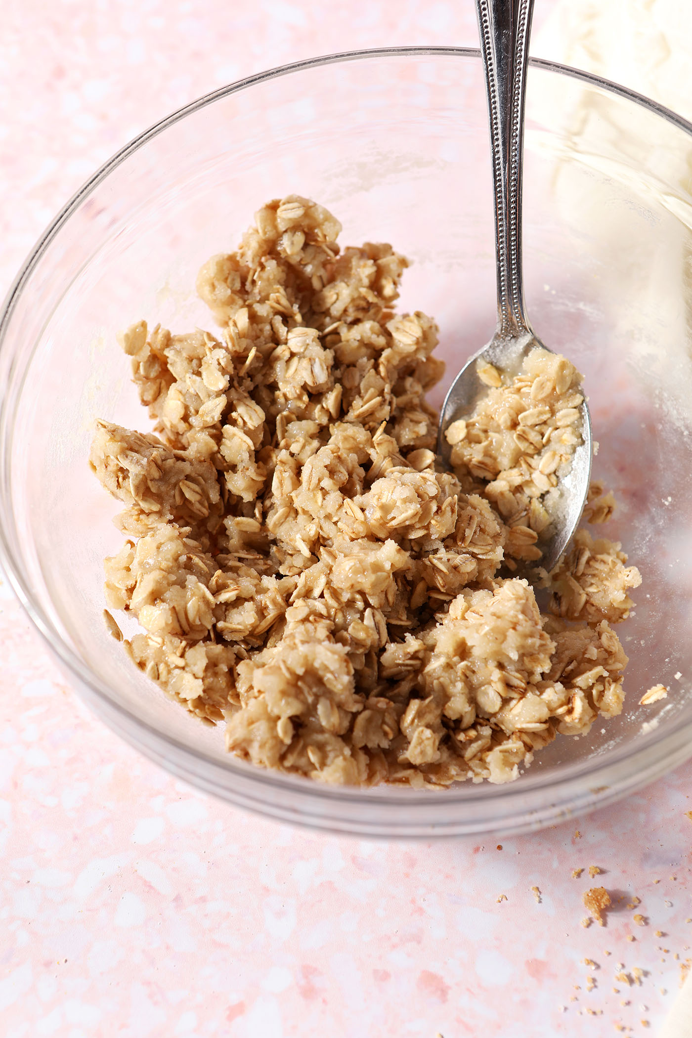 The crumble topping in a glass bowl with a spoon on a pink surface