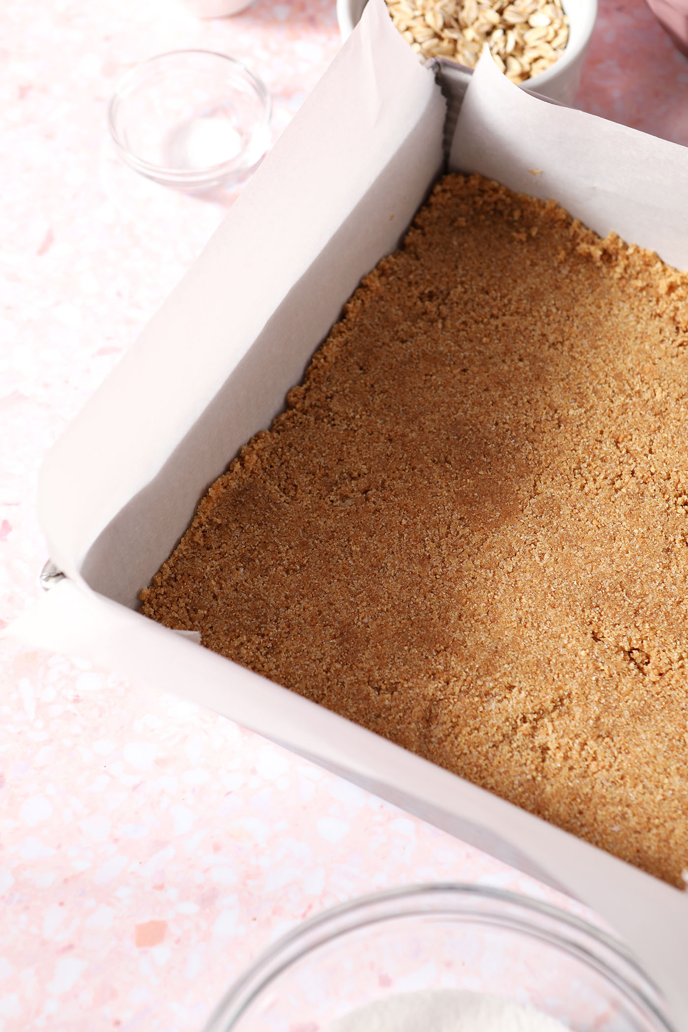 A graham cracker crumb crust in a square baking dish on a pink surface