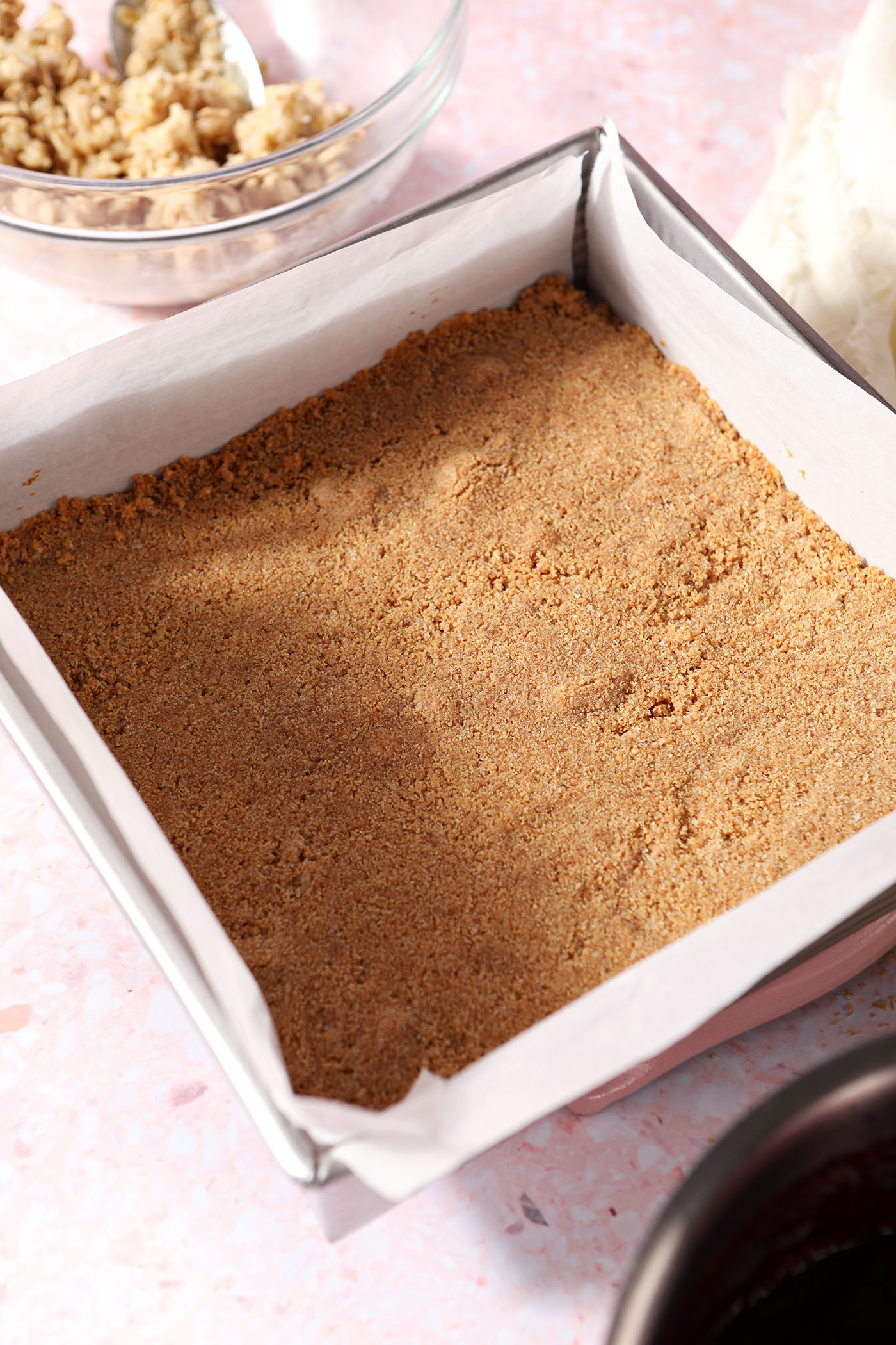 A baked graham cracker crust in a square baking pan surrounded by other ingredients on a pink surface