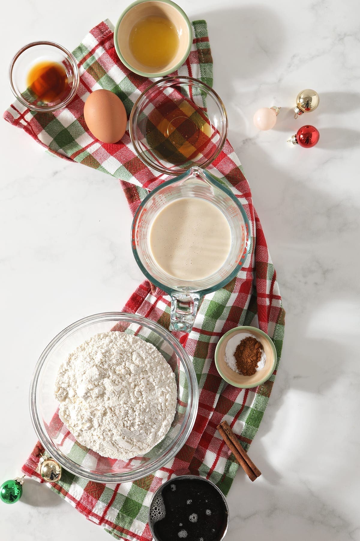 Ingredients for eggnog pancakes and the cinnamon syrup sit on top of a green and red plaid towel on marble next to small shiny ornaments