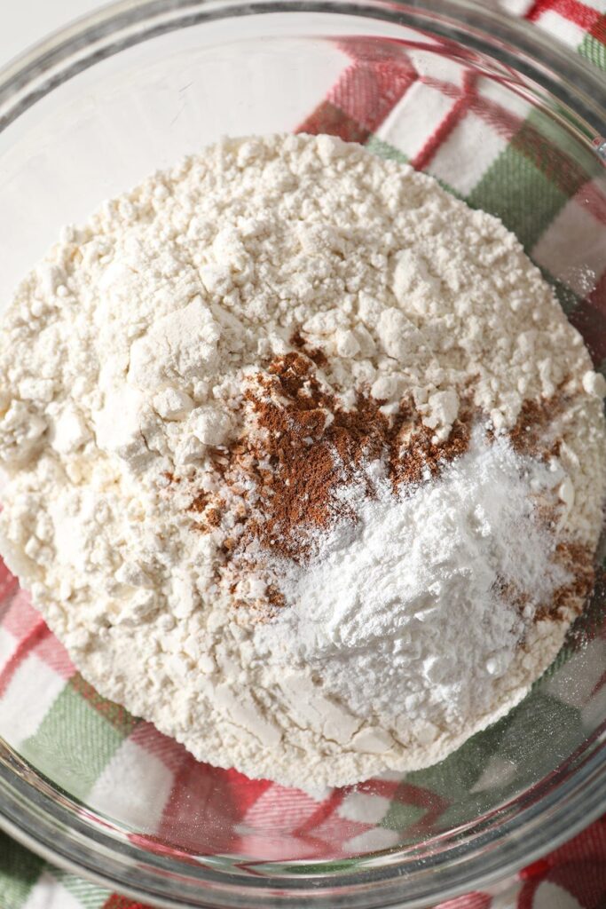 Flour, cinnamon and other dry ingredients in a clear glass bowl on top of a green and red plaid kitchen towel