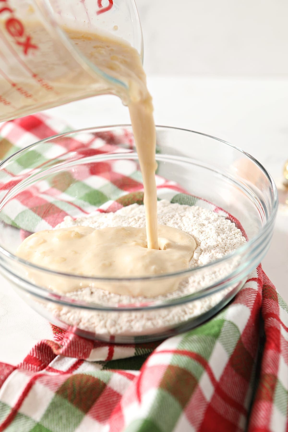 Wet ingredients pour into dry ingredients on top of a green and red plaid kitchen towel