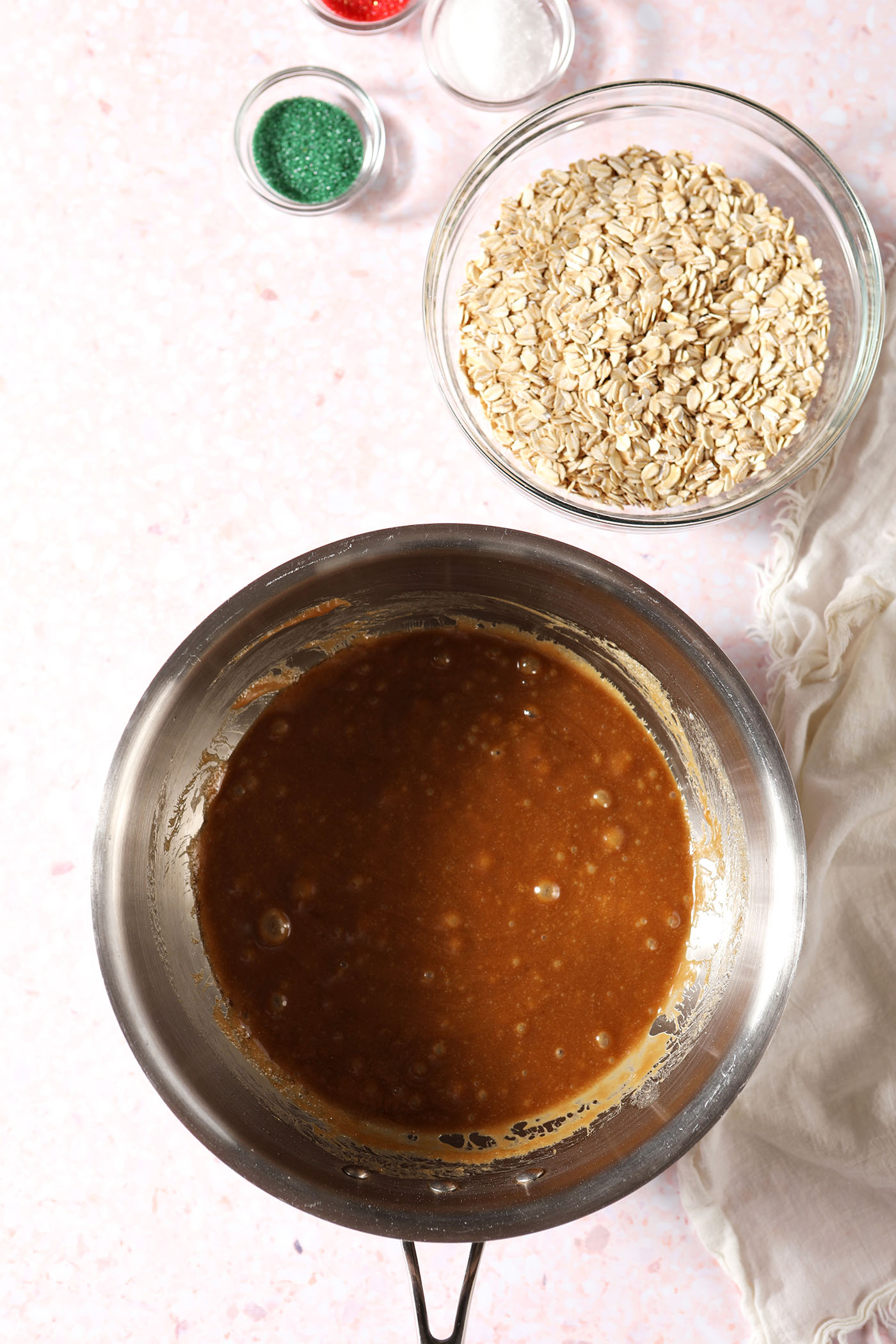 Melted no bake cookie ingredients in a saucepan next to a bowl of oats and colored sugars on a pink terrazzo surface