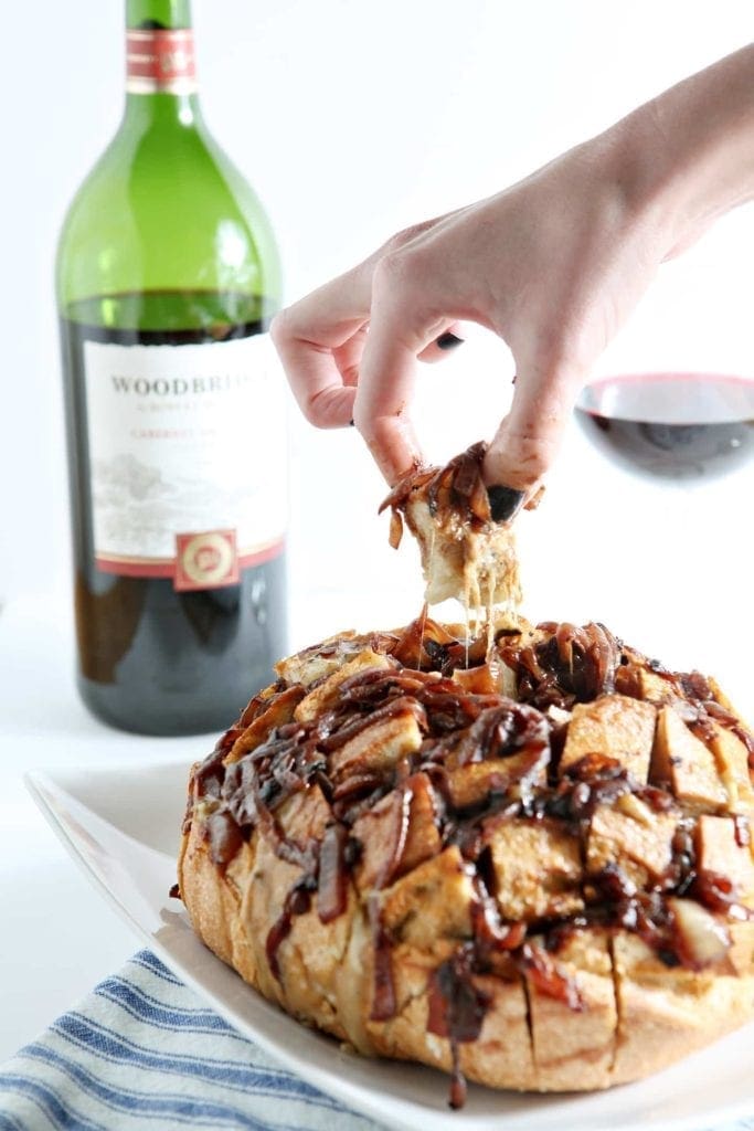 A person pulls a bite of Cheesy French Onion Pull-Apart Bread out of the boulle with a bottle of wine and a glass in the background