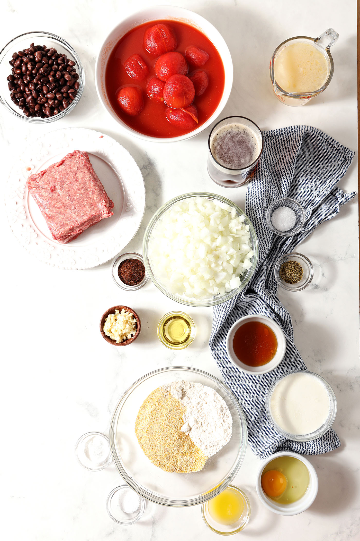 Ingredients to make chili and cornbread casserole in bowls on marble with a blue white striped linen