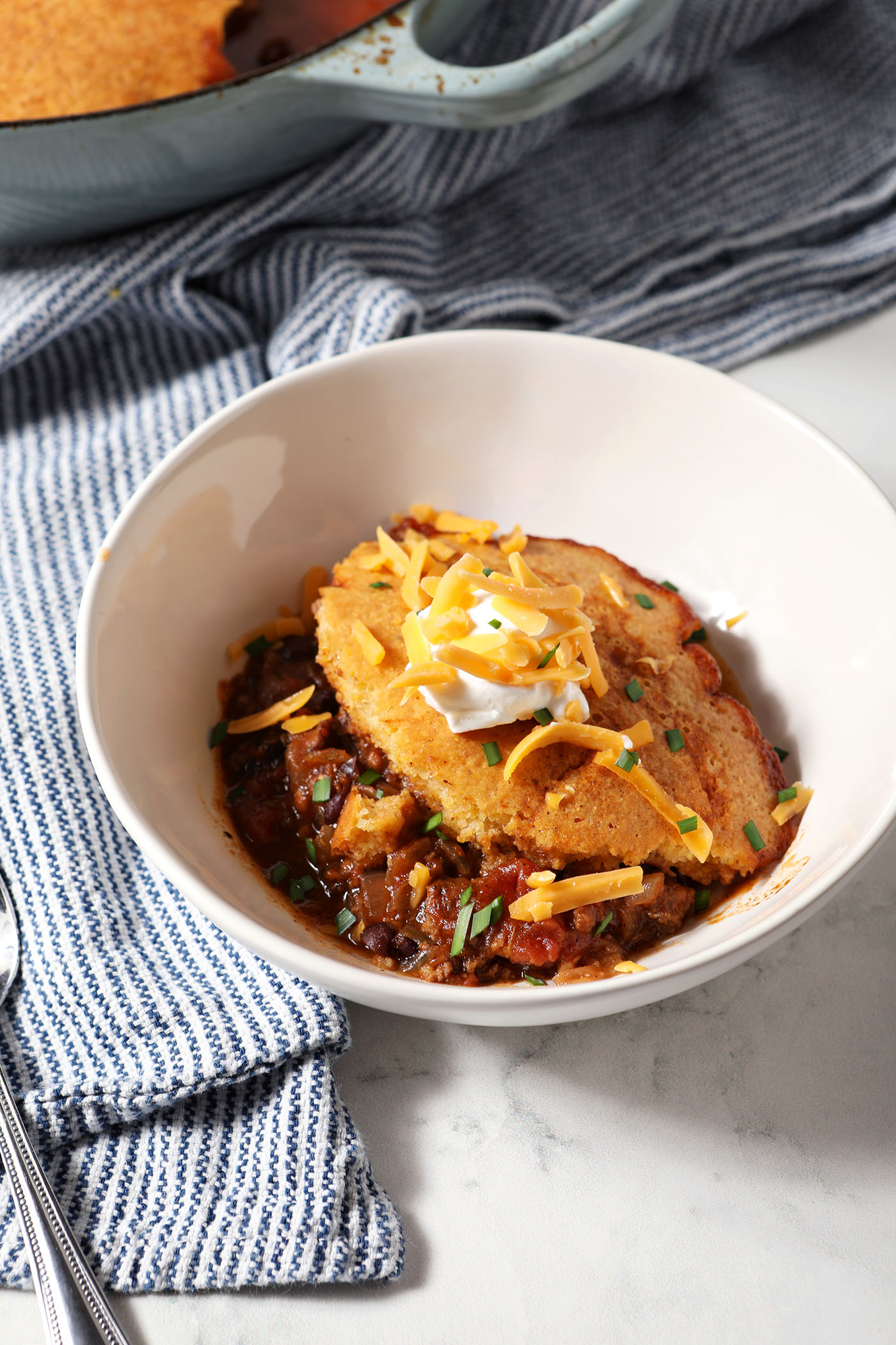 A bowl of Chili Cornbread Casserole topped with sour cream, shredded cheddar and chopped chives on marble next to a blue and white linen