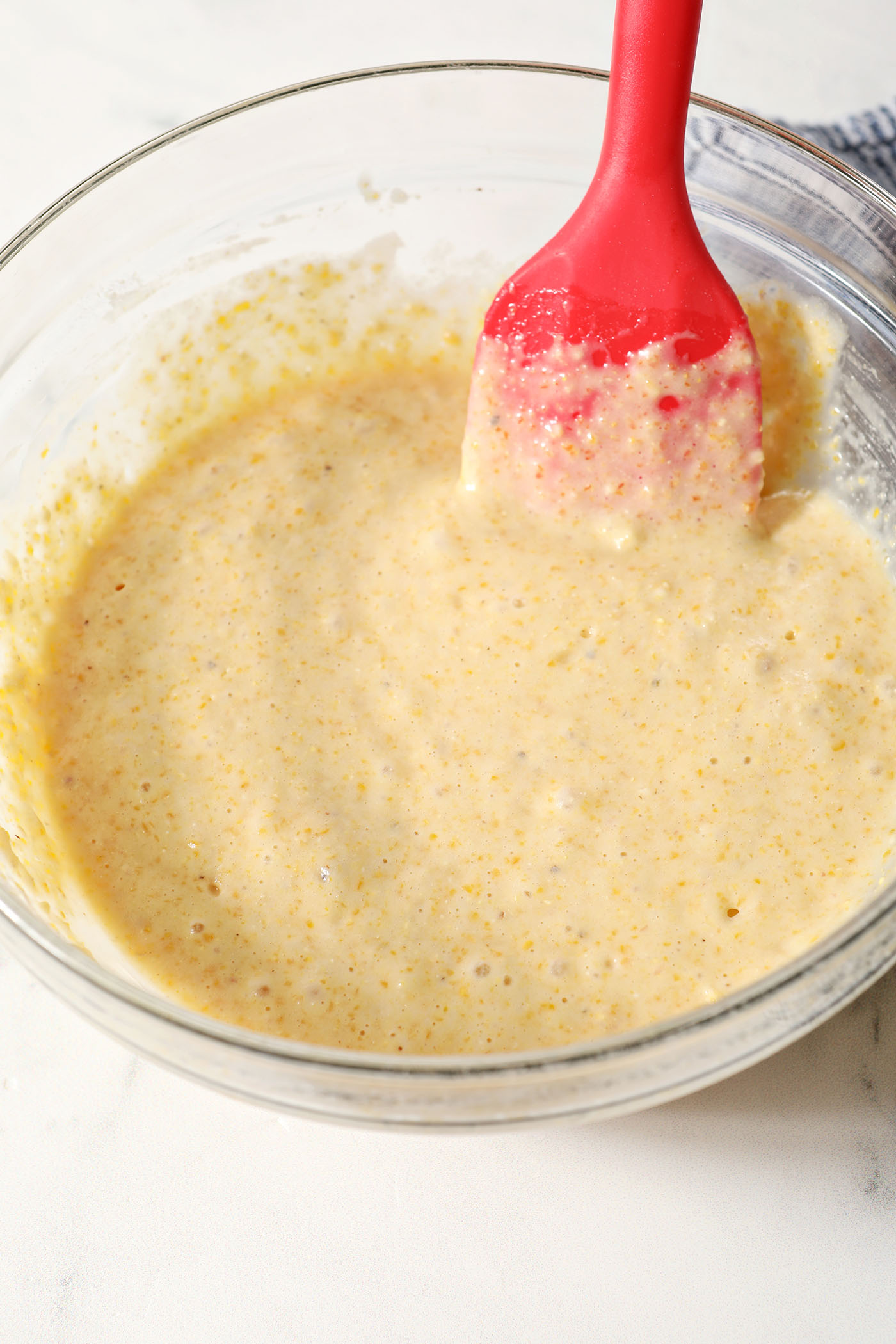Homemade cornbread batter in a glass bowl on a marble surface with a red spatula