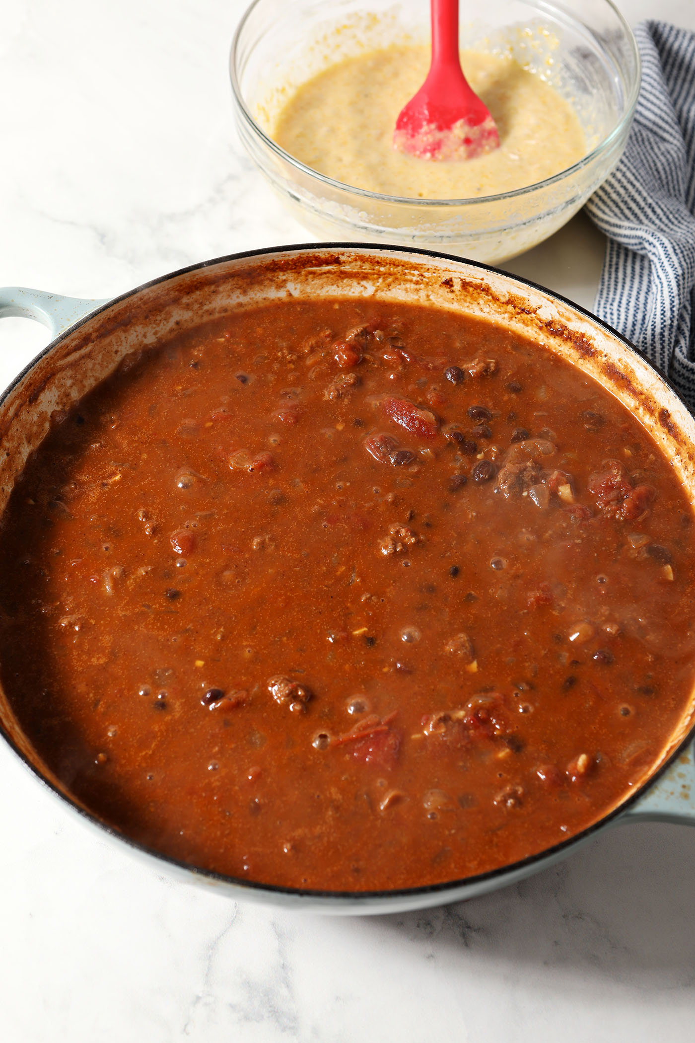 Skillet chili on a marble surface next to a bowl of cornbread batter and a blue and white linen