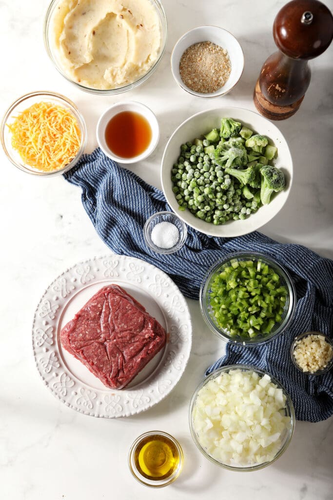 Ingredients to make cottage pie with ground beef in bowls and plates with a blue linen on marble