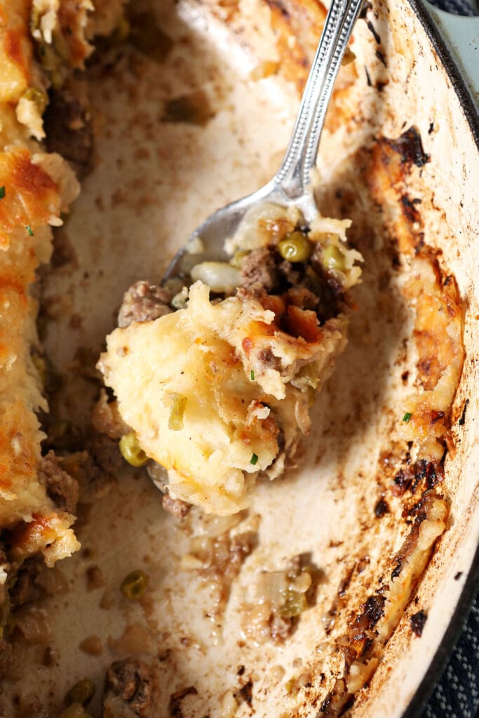 A large spoon holds a scoop of cottage pie from a large round baking dish