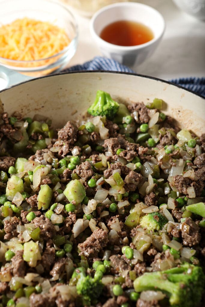 Cooked beef and veggies, including broccoli and peas, in a skillet next to a bowl of cheese