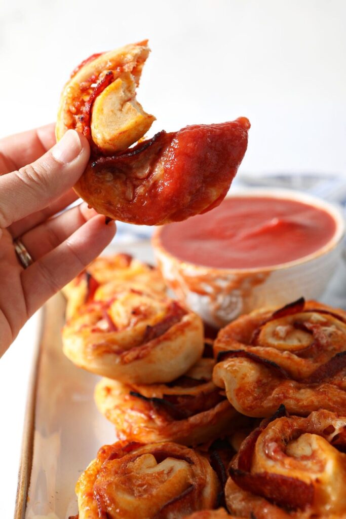 A bitten-into pizza roll is held above a platter with more rolls and marinara