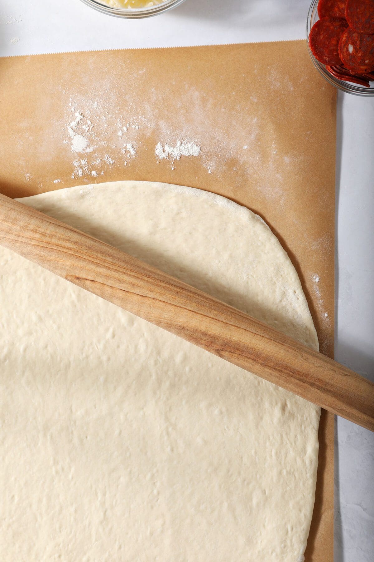 A wooden rolling pin rolls out pizza dough on parchment