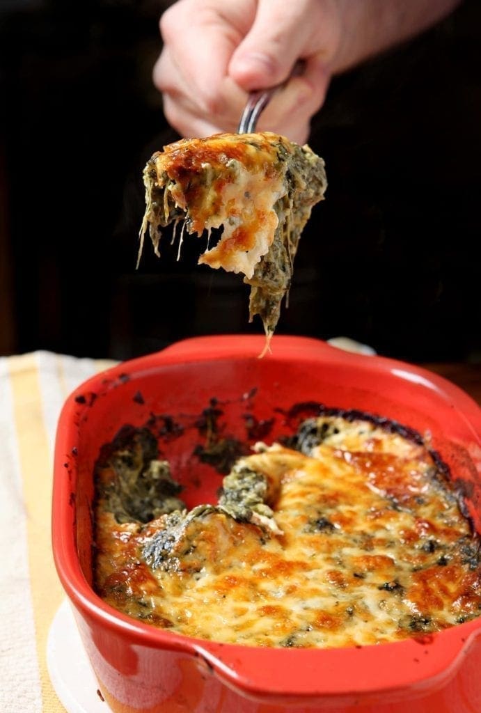 A spoonful of Spinach Madeline is lifted out of a red casserole dish