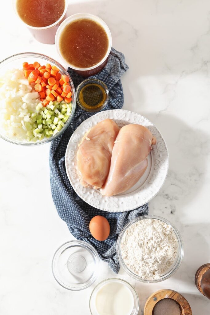 Chicken breasts, vegetables and other soup ingredients on a blue towel on a marble countertop