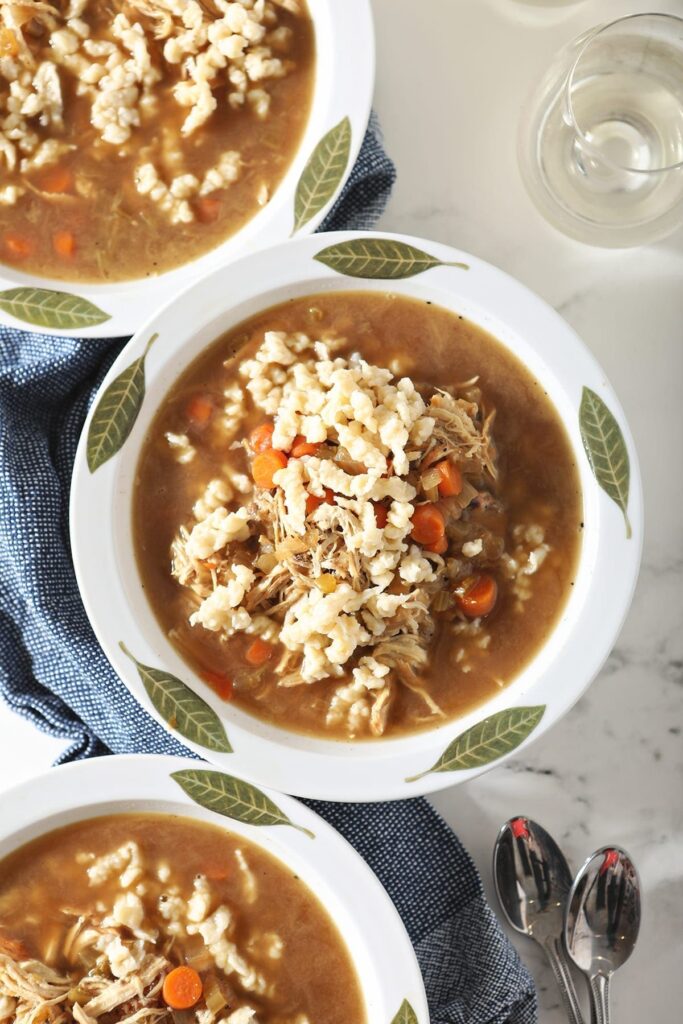 Three bowls of chicken soup with homemade German noodles sit on top of a blue towel next to spoons and glasses of wine