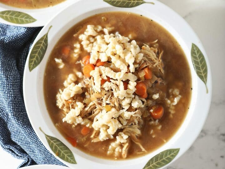 Three bowls of chicken soup with homemade German noodles sit on top of a blue towel next to spoons and glasses of wine