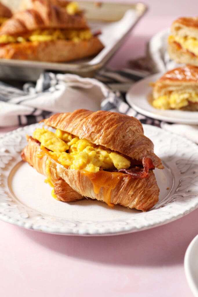 a breakfast croissant sandwich on a white plate on a pink surface next to more plates of more breakfast sandwiches