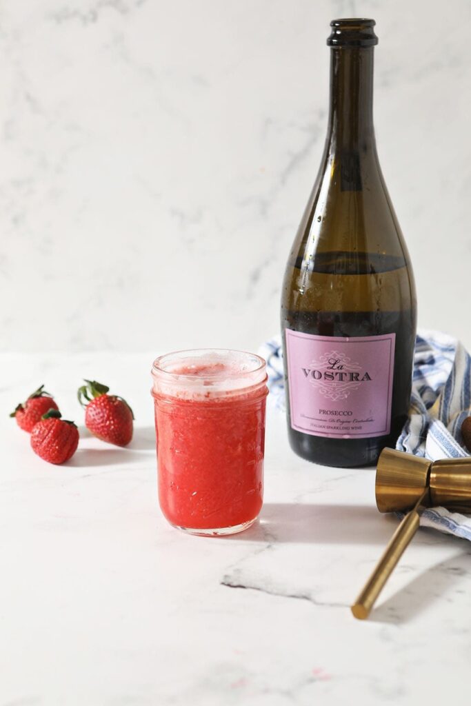 A jar of strawberry simple syrup sits next to a bottle of champagne, strawberries and a gold jigger