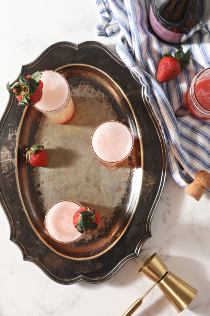 Three Strawberry Champagne mixed drinks sit on a metal tray next to ingredients and a gold jigger