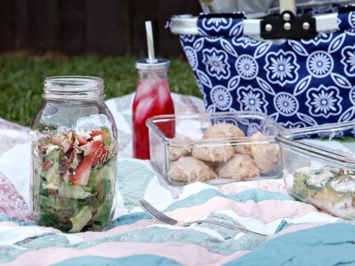 Containers of food and drink on blanket next to blue picnic basket