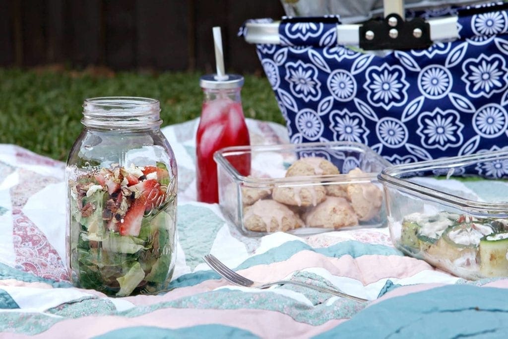 Containers of food and drink on blanket next to blue picnic basket 