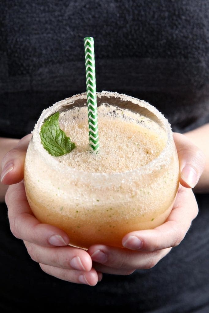 A woman holds a Cantaloupe Mint Margaritas, garnished with a sprig of mint and rimmed with sugar, before drinking