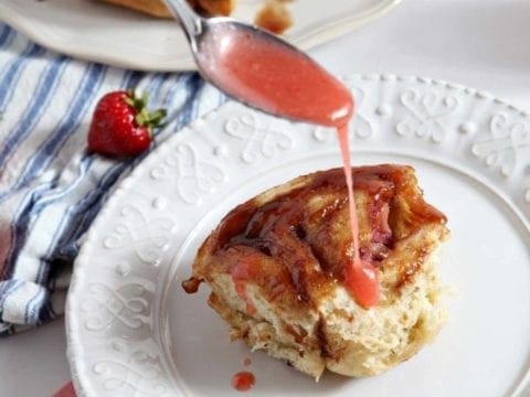 Spoon pouring glaze over strawberry cinnamon bun on white plate