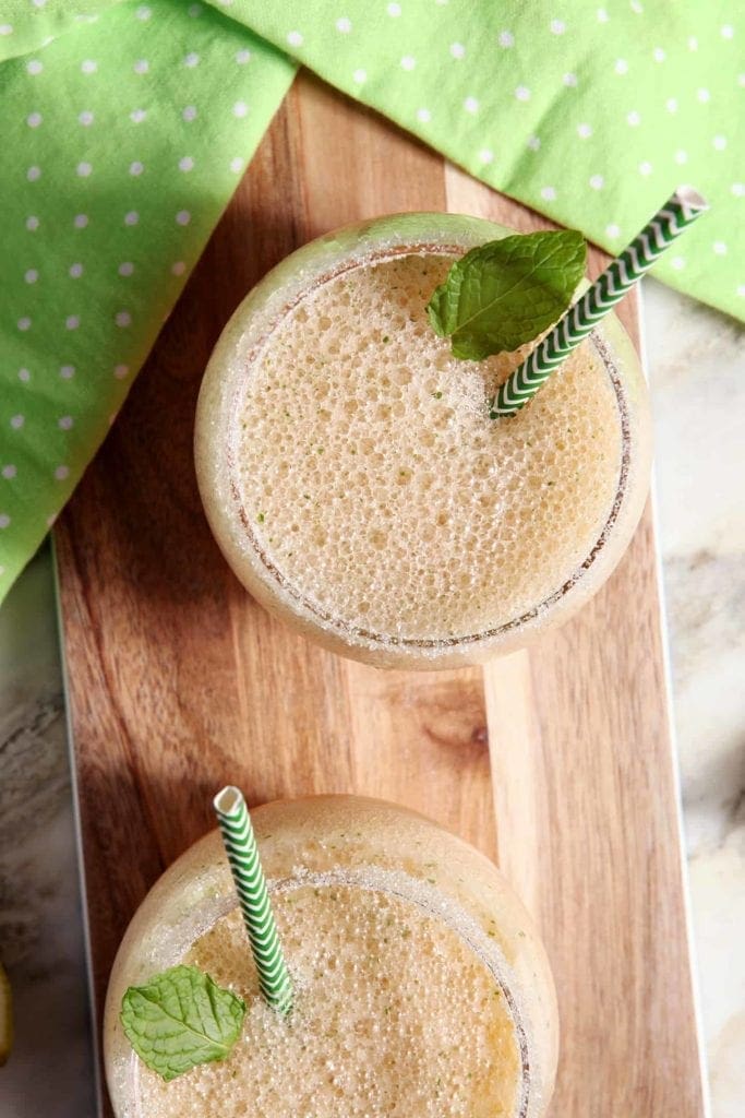 Overhead image of two melon Margaritas are served on a wooden serving platter with lime wedges, rimmed with sugar, and garnished with mint