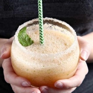 A woman holds a Cantaloupe Mint Margarita, rimmed with sugar and garnished with a sprig of mint, in hand before drinking