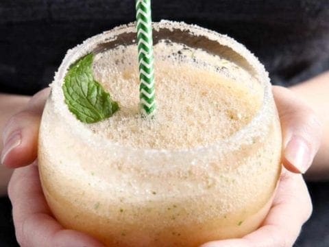 A woman holds a Cantaloupe Mint Margarita, rimmed with sugar and garnished with a sprig of mint, in hand before drinking