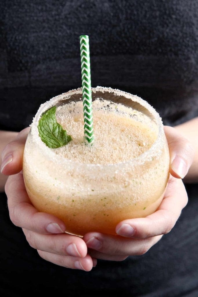 A woman holds a Cantaloupe Mint Margarita, rimmed with sugar and garnished with a sprig of mint, in hand before drinking