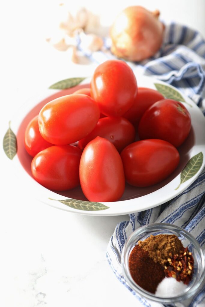 Tomatoes in a bowl next to spices, an onion and garlic on marble