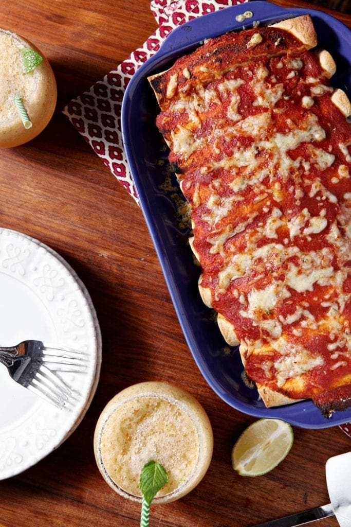 Overhead shot of a baking dish full of baked enchiladas, surrounded by plates and margaritas, on a wooden background