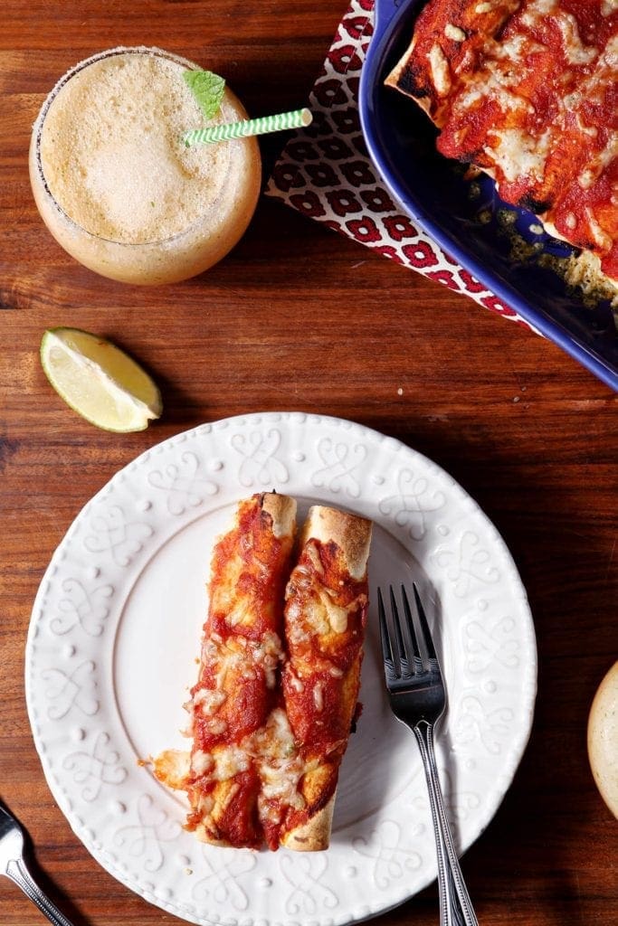 Two Pork Carnitas Enchiladas, served on a white plate with a fork, surrounded by lime wedges and the platter of enchiladas.