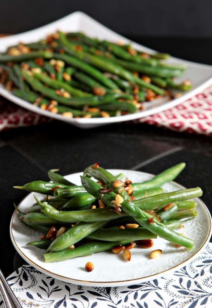 A plate of steamed green beans with toasted pine nuts and shallots on a dark background with a platter of green beans behind it