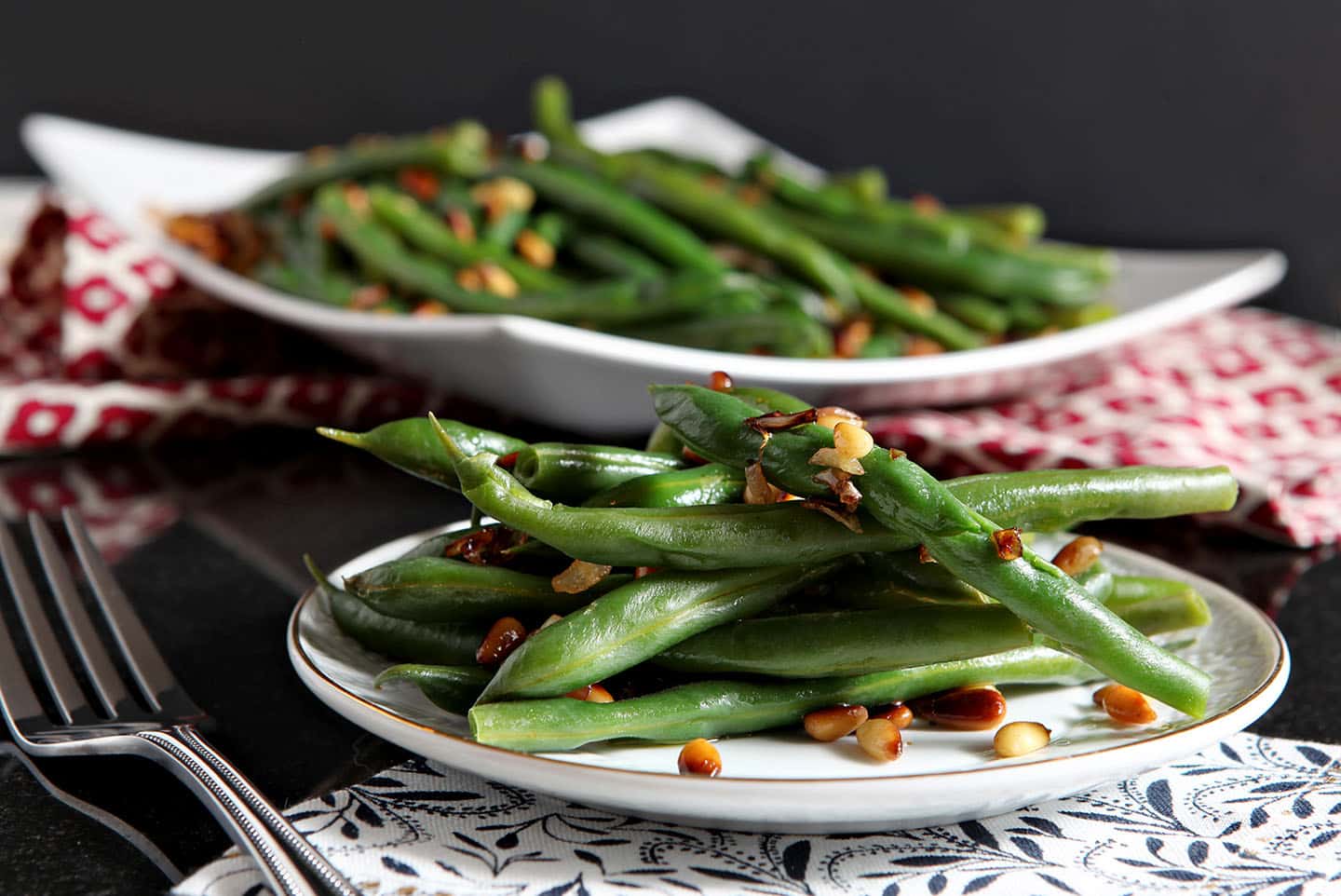 A white plate holds a serving of vegan green beans with pine nuts on a dark background