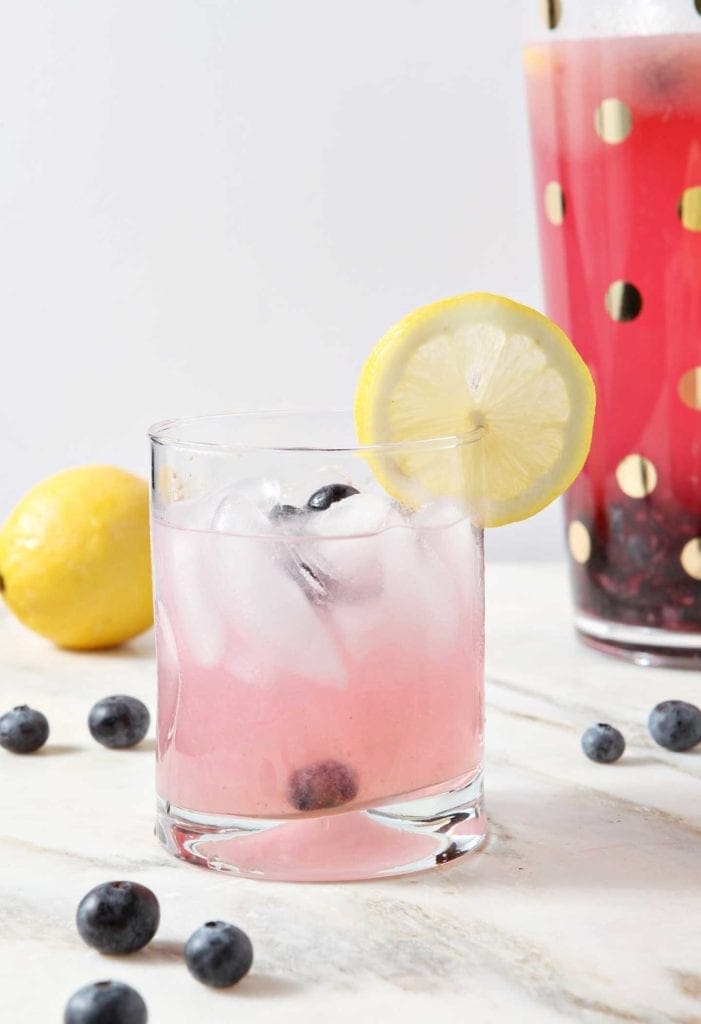 A glass of homemade blueberry lemonade with a lemon slice in front of a pitcher