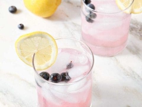 Two glasses of Blueberry Lemonade, from the side, garnished with fresh blueberries and wheels of lemons