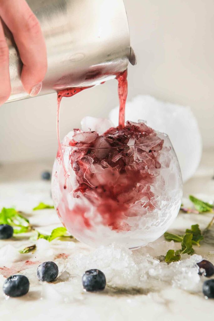 A Blueberry Mint Julep pours into a glass of ice from a cocktail shaker