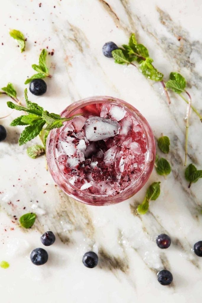 A Blueberry Mint Julep sits on marble surrounded by blueberries and mint leaves