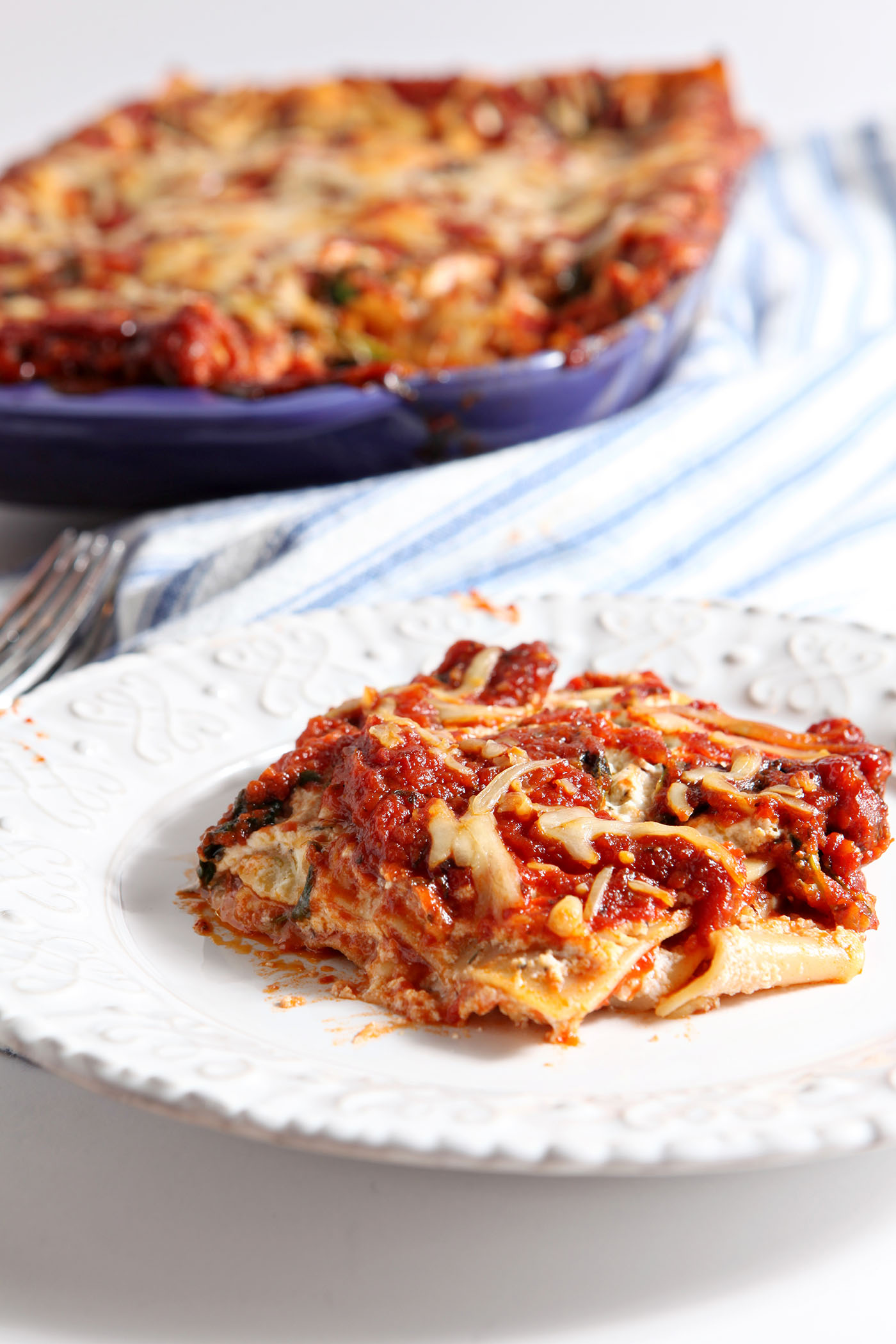 A large slice of homemade spinach lasagna on a white plate in front of a purple casserole with more lasagna