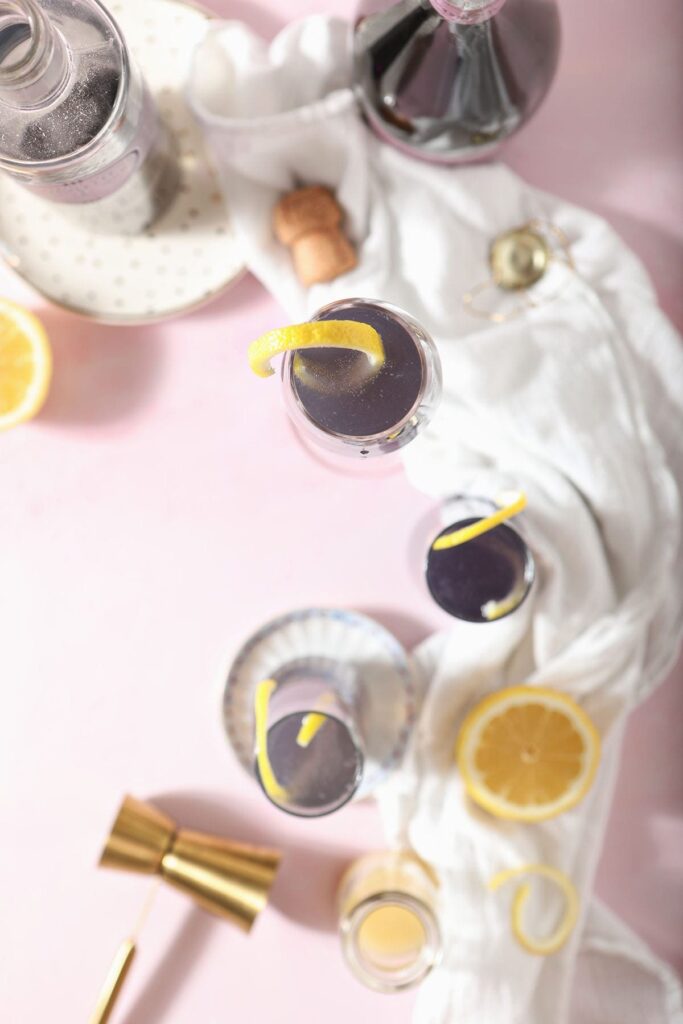 Several creme de violette cocktails on a pink surface with bottles, lemons and cocktail tools