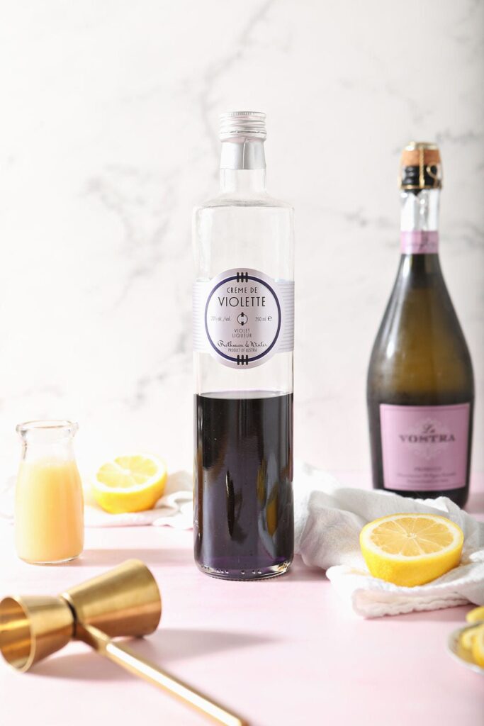 A bottle of creme de violette sits next to lemon juice, champagne and lemons