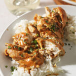 Sliced mustard chicken on top of rice on a white plate next to a glass of wine