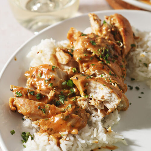 Sliced mustard chicken on top of rice on a white plate next to a glass of wine